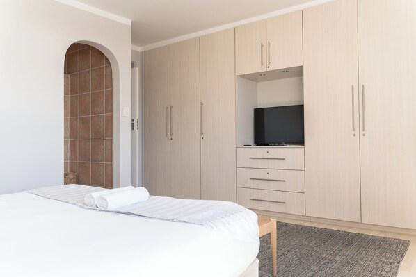 Une chambre à coucher avec un lit, un téléviseur posé sur une étagère et une armoire avec des tiroirs et un espace pour suspendre les vêtements.