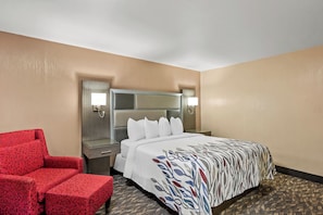 A hotel room with a bed, a red armchair, a nightstand, and a wall-mounted lamp. - Red Roof Inn Yuba City Bonanza - Marysville (Yuba City)