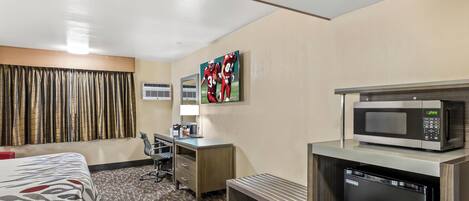 Habitación de hotel con cama, escritorio, microondas, lavavajillas, banco, televisor y ventana con cortinas.