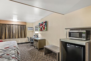 A hotel room with a bed, a desk, a microwave, a dishwasher, a bench, a TV, and a window with curtains.