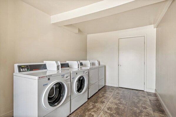 A laundry room with three front-loading washing machines and a dryer, a white door, and a tiled floor. - Red Roof Inn Yuba City Bonanza - Marysville (Yuba City)