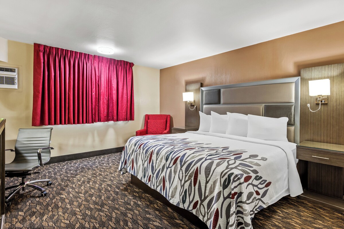 Una habitación de hotel con una cama grande, una silla roja, un escritorio con silla y una lámpara de pared.