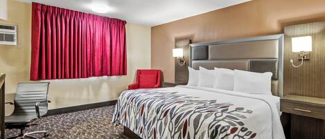 A hotel room with a large bed, a red chair, a desk with a chair, and a wall-mounted lamp.