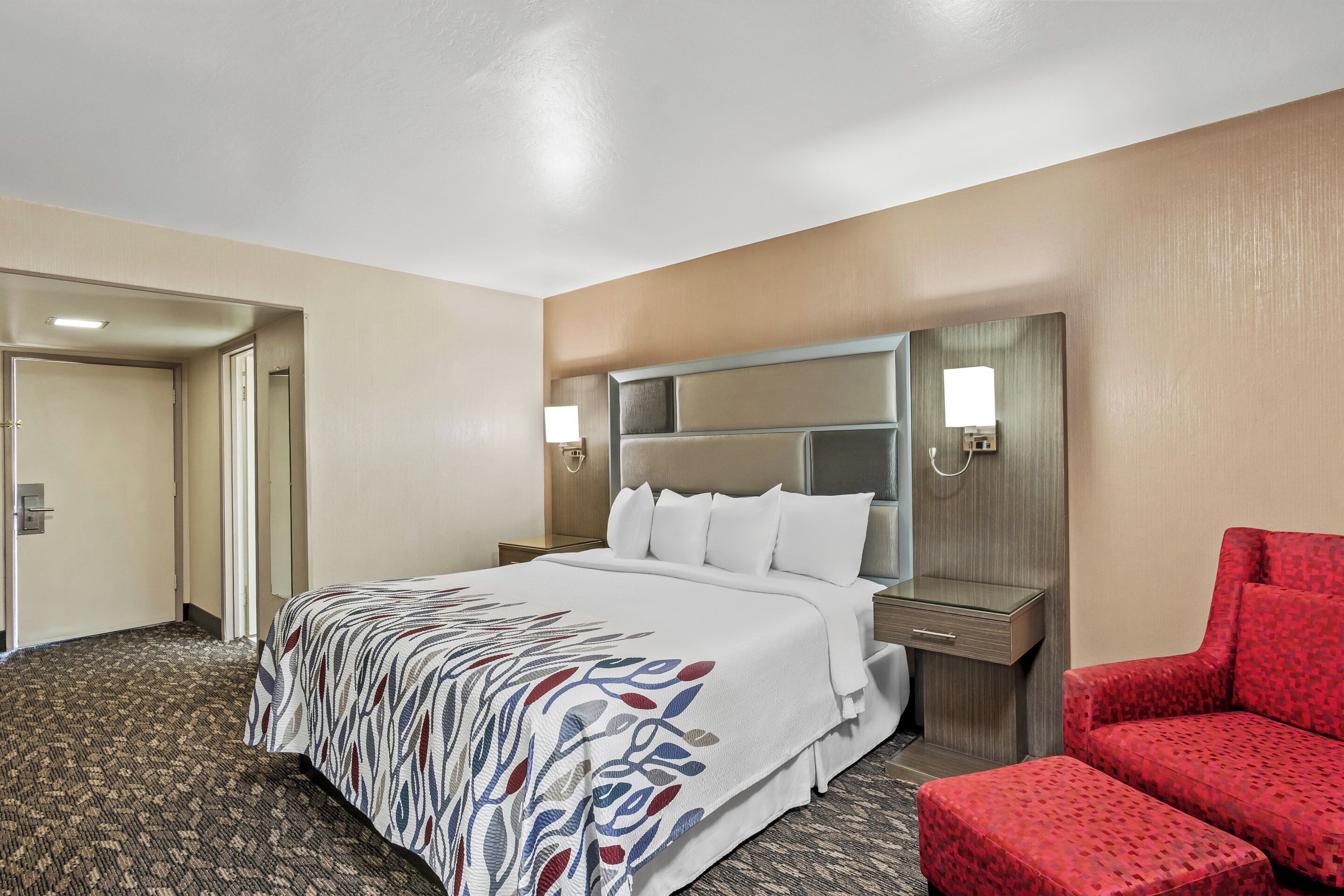 Una habitación de hotel con una cama, un sillón rojo, una mesita de noche y un espejo.