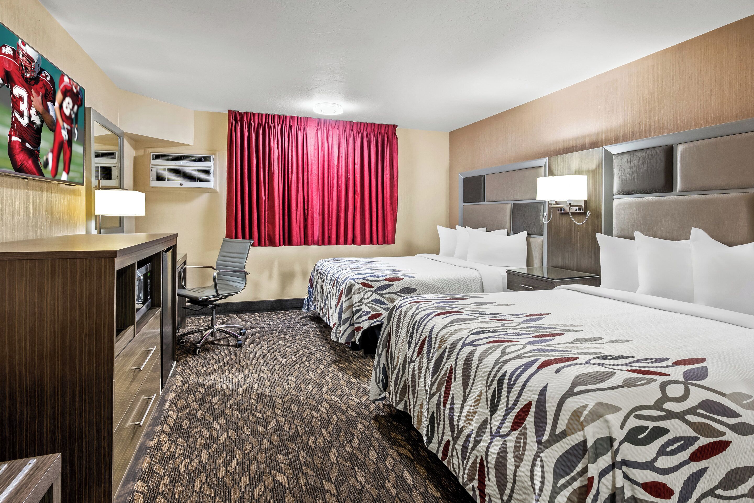 A hotel room with two beds, a desk, a chair, a large mirror, and a red curtain.