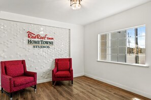Una habitación con pared de diseño geométrico, dos sillones rojos, un ventanal con vistas al exterior y un letrero que dice 'RedRoof Inn HOME TOWN STUDIOS'.