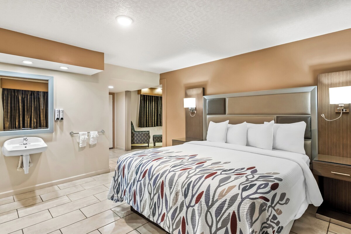 Habitación de hotel con cama grande, mesita de noche, lámpara de pared, espejo y baño con lavamanos y toallero.