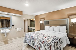 Habitación de hotel con cama grande, mesita de noche, lámpara de pared, espejo y baño con lavamanos y toallero.