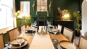 Un espace repas avec une longue table dressée pour six personnes, agrémenté d’un élément décoratif central, d’un miroir au-dessus de la cheminée et d’un mur vert.