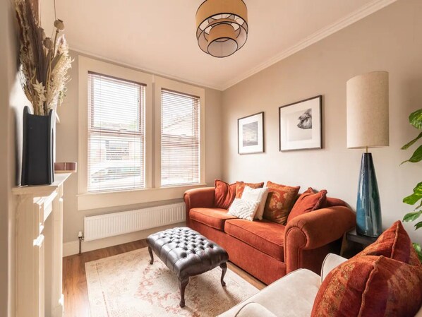 A living room with a sofa, ottoman, and a lamp. There are two windows with blinds, a radiator, and a fireplace.