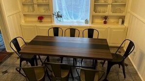 A dining area with a wooden table and six chairs, a window with a sheer curtain, and shelves with glassware and decorative items. - Refined Home w/Game Room & Large Lounge (Westworth Village)