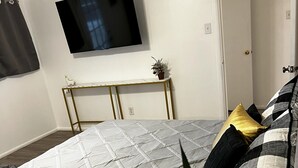 A bedroom with a bed, a TV mounted on the wall, a small table with a plant, and a door. - Refined Home w/Game Room & Large Lounge (Westworth Village)
