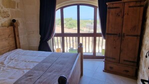 A bedroom with a large bed, a wooden wardrobe, and a window with a view of greenery and a clear sky. - The old Mill (Xaghra)