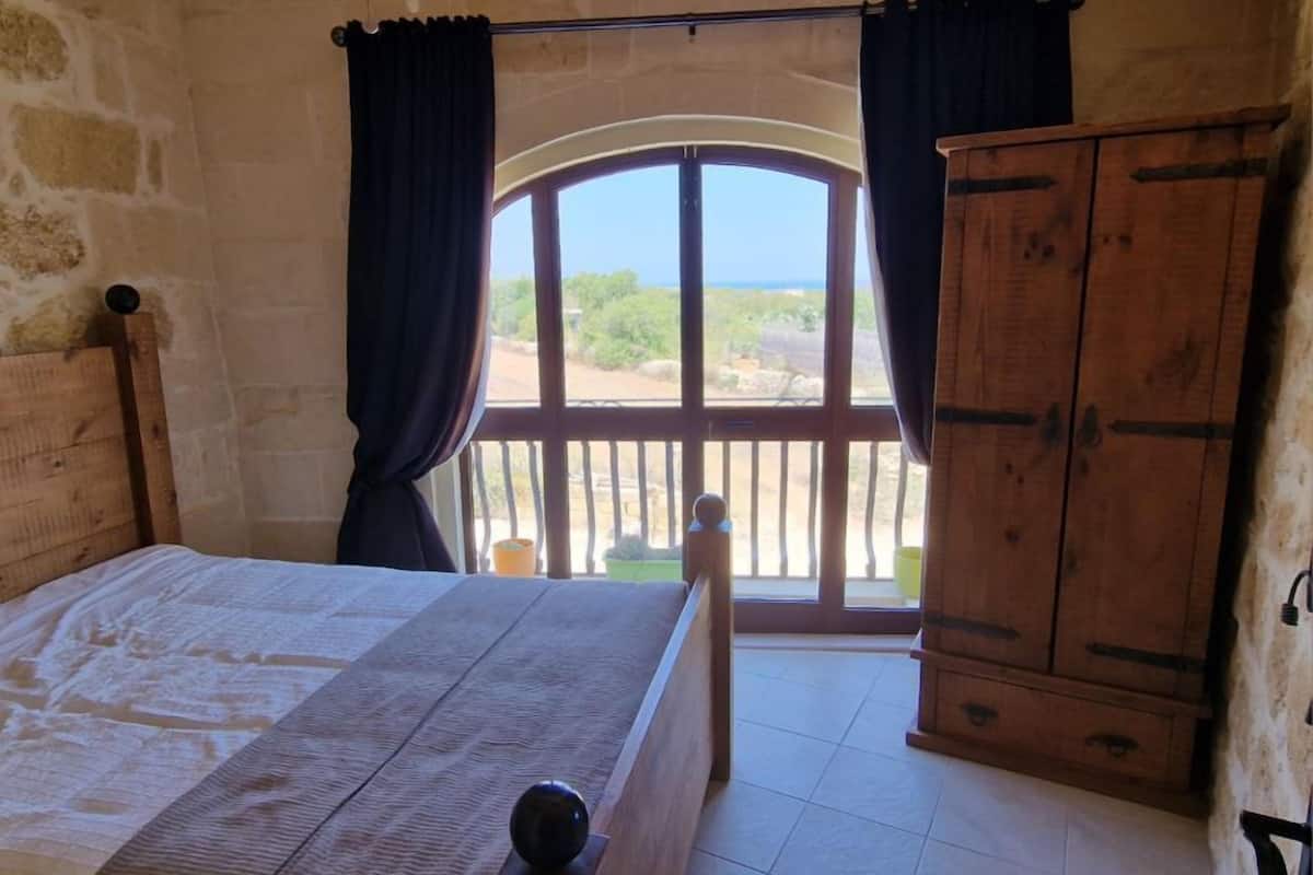 A bedroom with a large bed, a wooden wardrobe, and a window with a view of greenery and a clear sky.