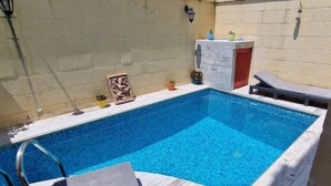 A rectangular swimming pool with a tiled edge, a lounge chair, a small table, and a wall-mounted light fixture. - The old Mill (Xaghra)