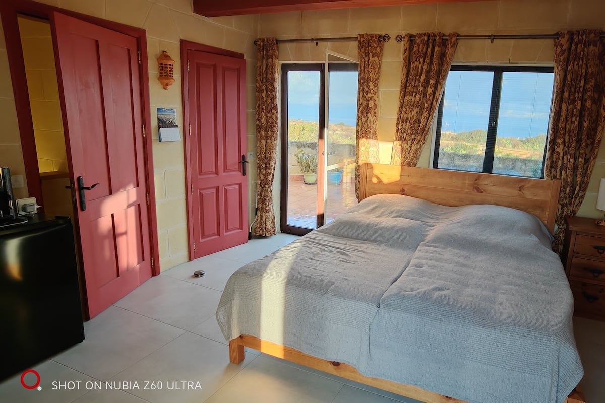 A bedroom with a bed, a wooden nightstand, a red door, and a view of the outdoors.