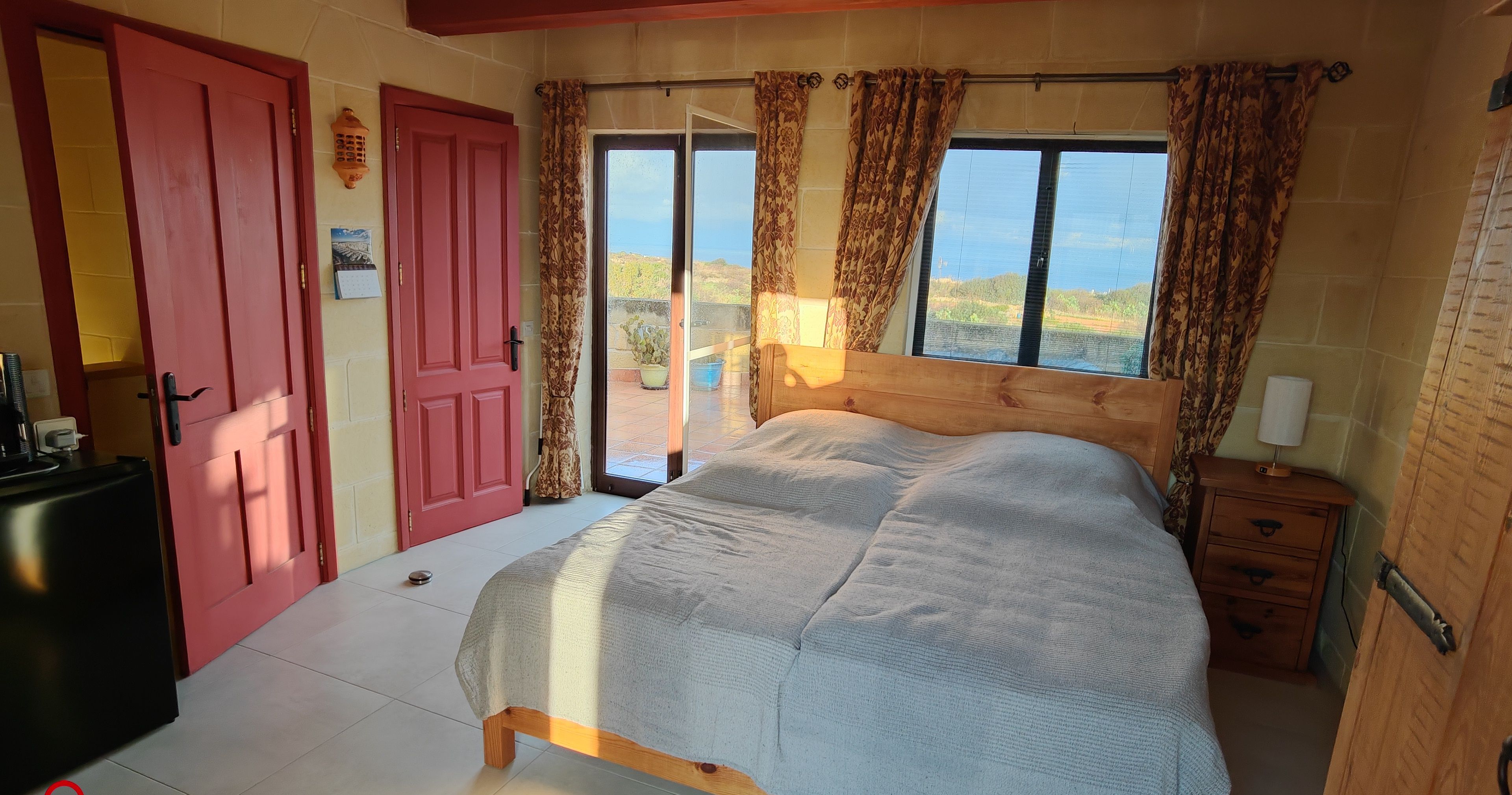 A bedroom with a bed, a wooden nightstand, a red door, and a view of the outdoors.