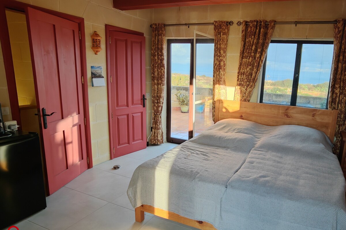 A bedroom with a bed, a wooden nightstand, a red door, and a view of the outdoors.