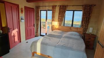 A bedroom with a bed, a wooden nightstand, a red door, and a view of the outdoors.