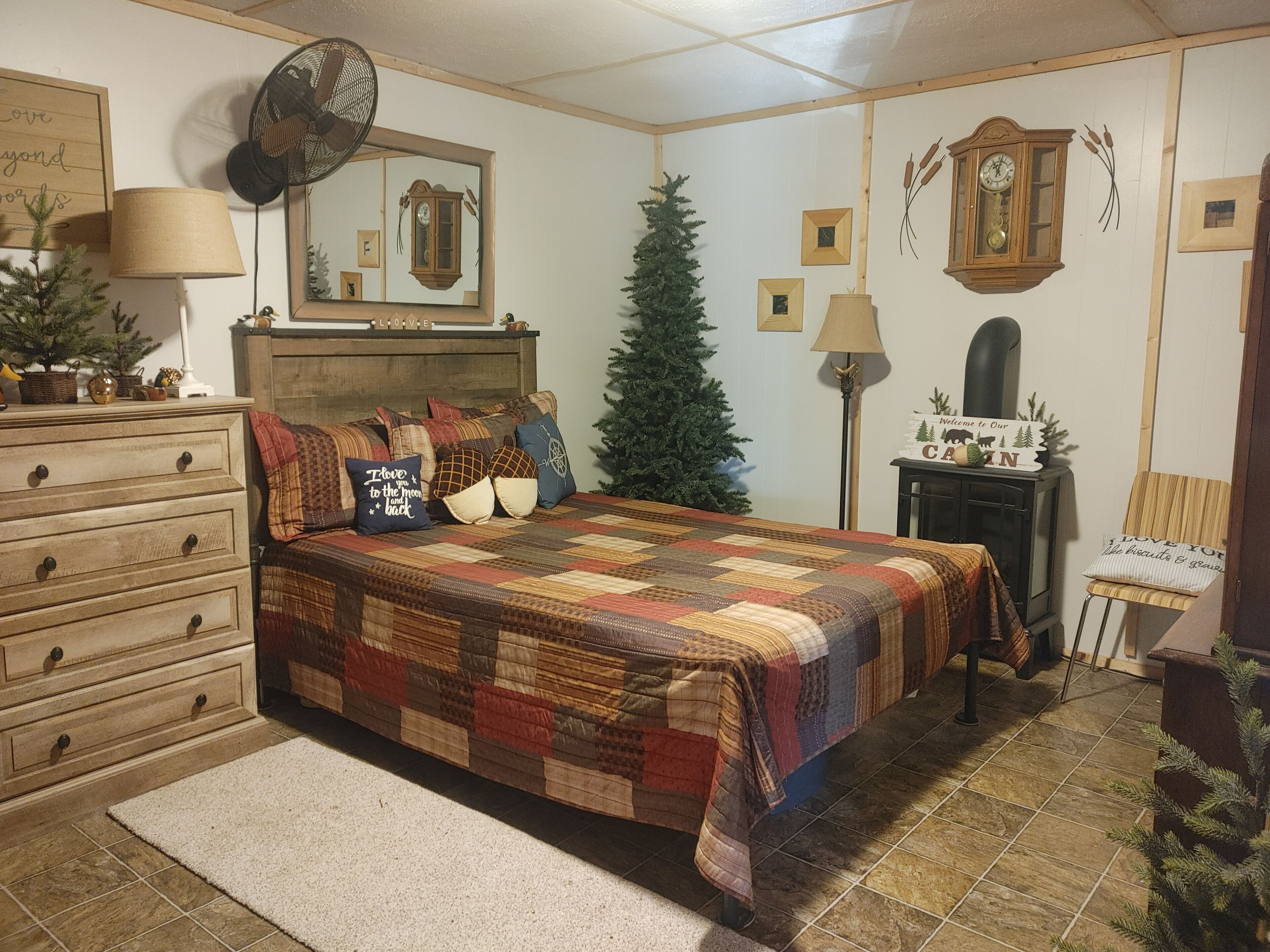 Une chambre avec une commode en bois, un lit recouvert d’une courtepointe, une lampe sur pied, un petit sapin de Noël et une horloge au mur.