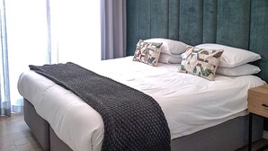 Ein modernes Schlafzimmer mit einem großen Bett, einem grau gepolsterten Kopfteil, weißer Bettwäsche und einer grauen Decke. An der Wand hängen zwei gerahmte Kunstwerke und ein Nachttisch steht neben dem Bett.