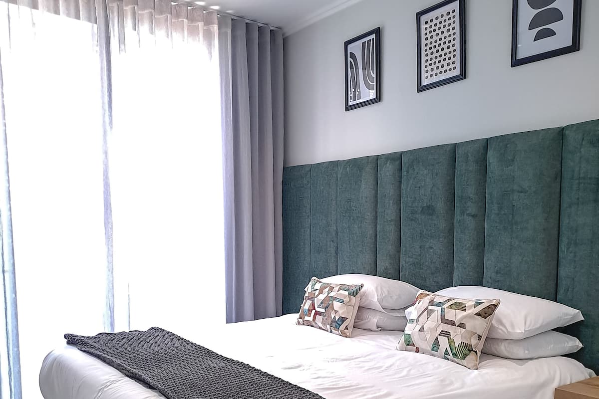 Ein modernes Schlafzimmer mit einem großen Bett, einem grau gepolsterten Kopfteil, weißer Bettwäsche und einer grauen Decke. An der Wand hängen zwei gerahmte Kunstwerke und ein Nachttisch steht neben dem Bett.