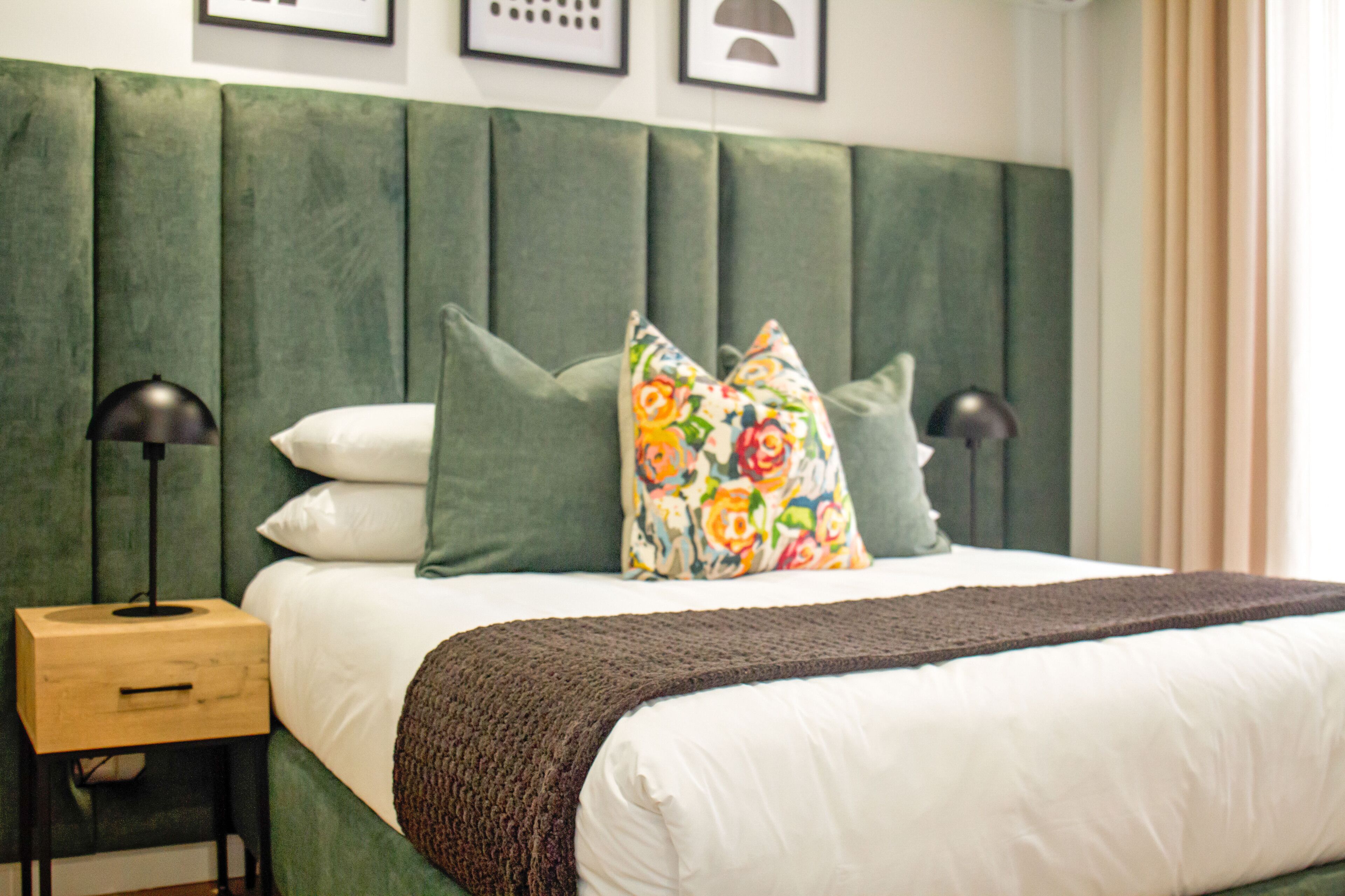 A neatly made bed with a green upholstered headboard, two bedside lamps, and a wooden nightstand.