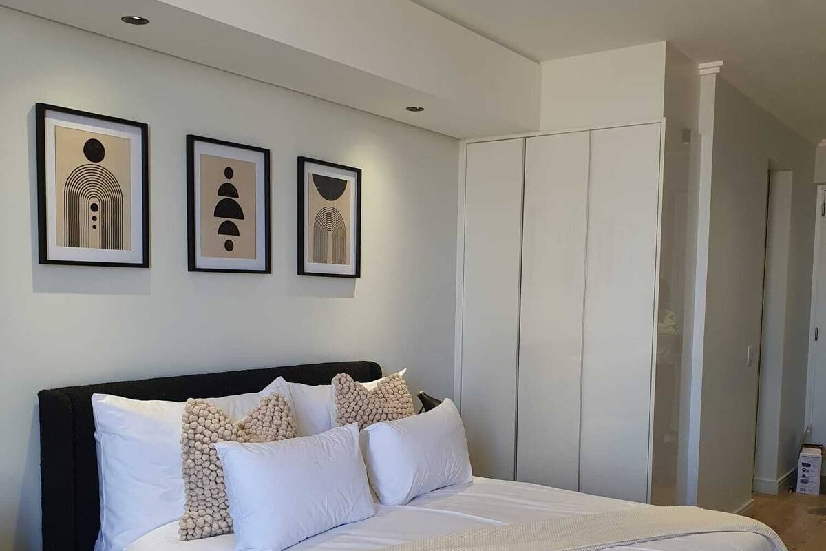 Ein modernes Schlafzimmer mit einem großen Bett, einem dunklen Kopfteil und drei gerahmten abstrakten Kunstwerken an der Wand.