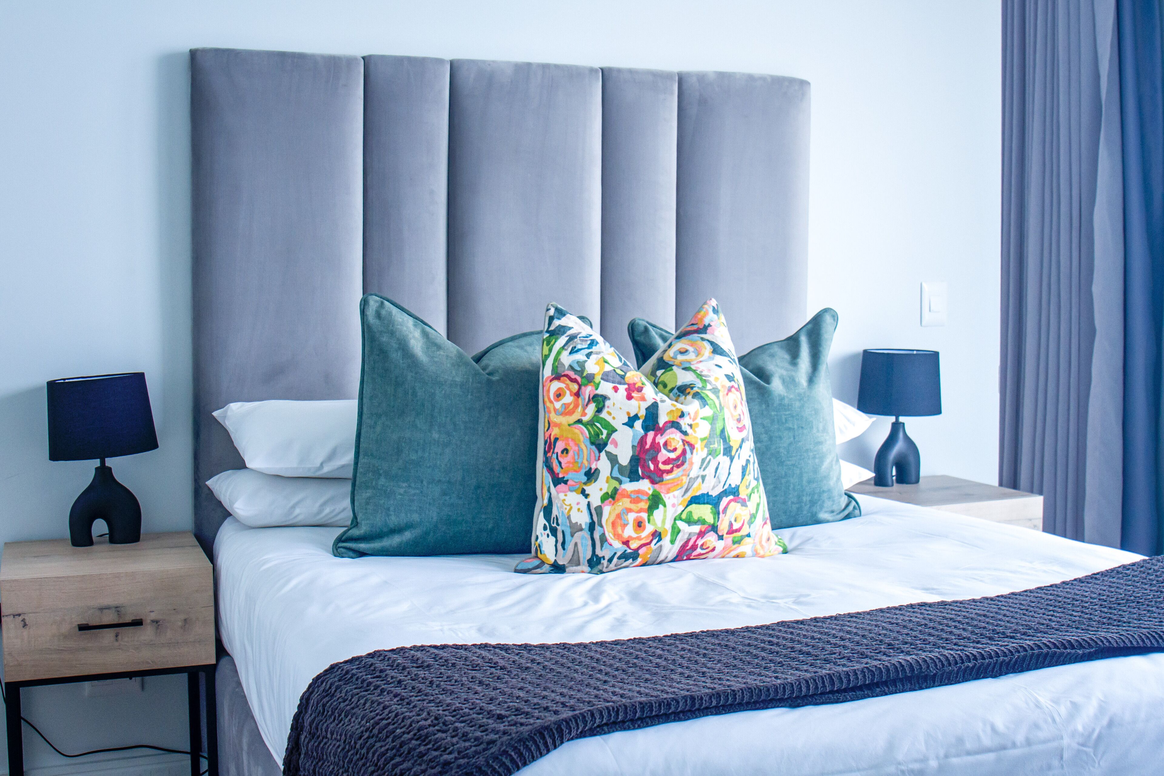 Una cama bien hecha con un cabecero tapizado en gris, dos mesitas de noche con lámparas y un cojín floral colorido.