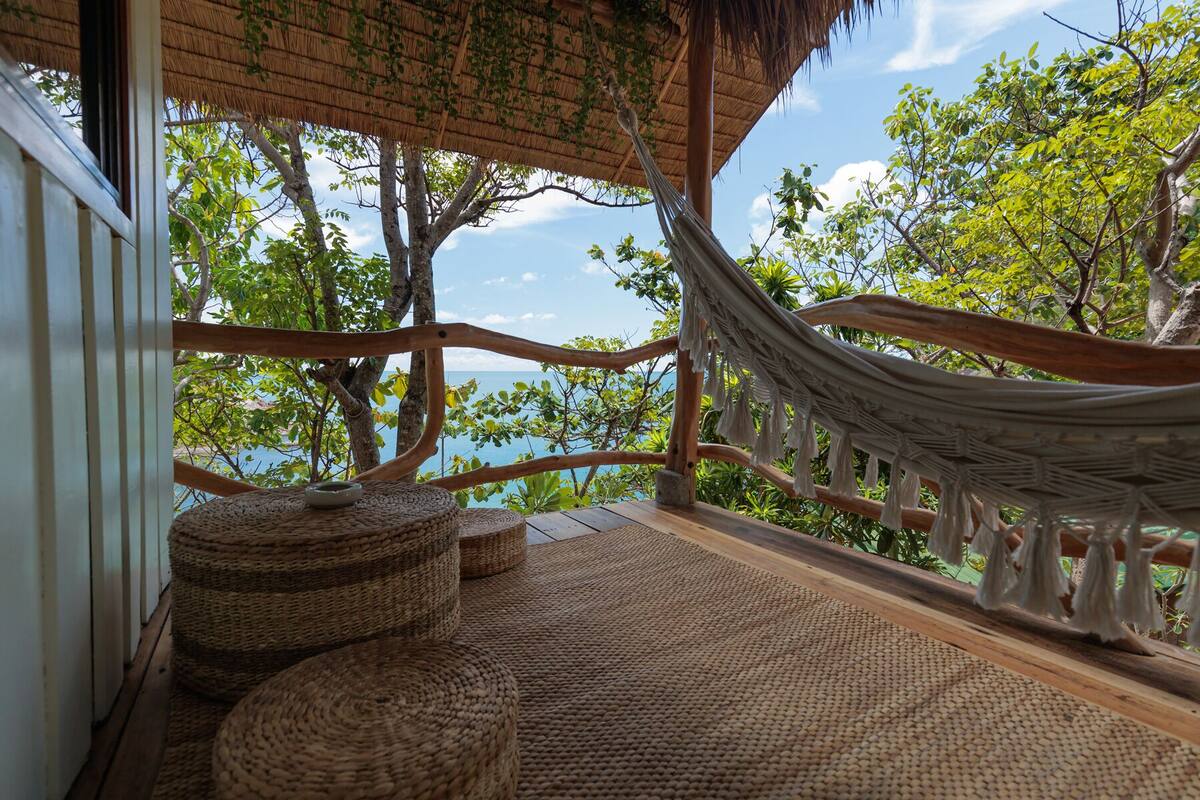 A hammock under a thatched roof, wicker furniture, and a view of trees and water.