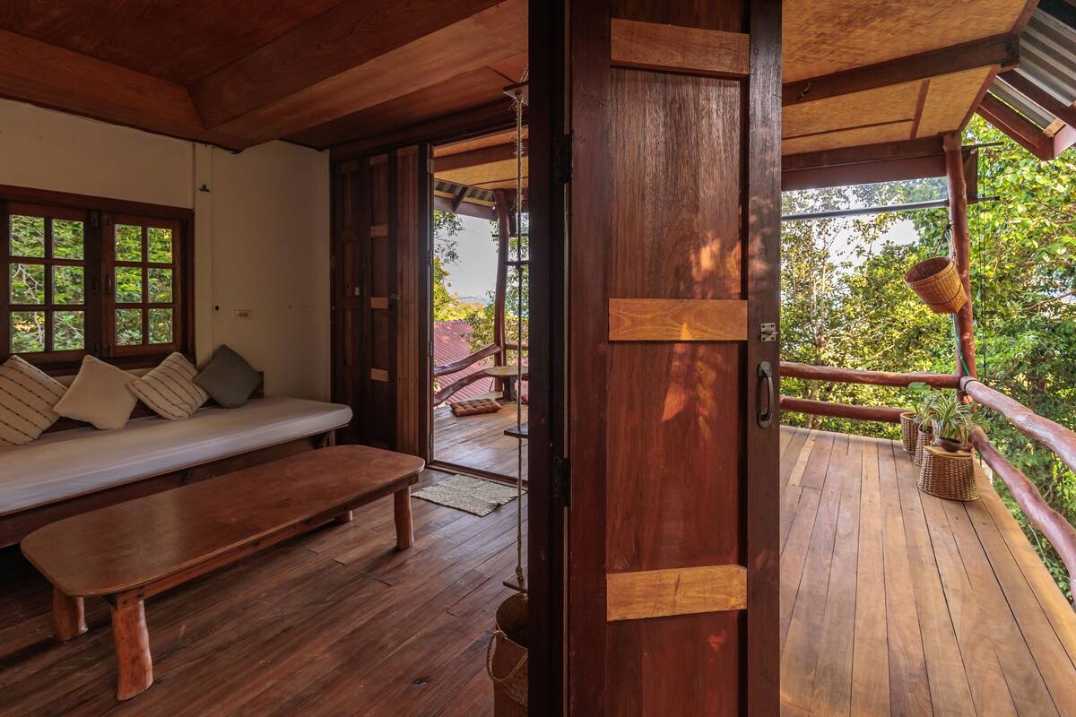A wooden interior with a bench and a sofa, a wooden door leading outside, and a balcony with potted plants.