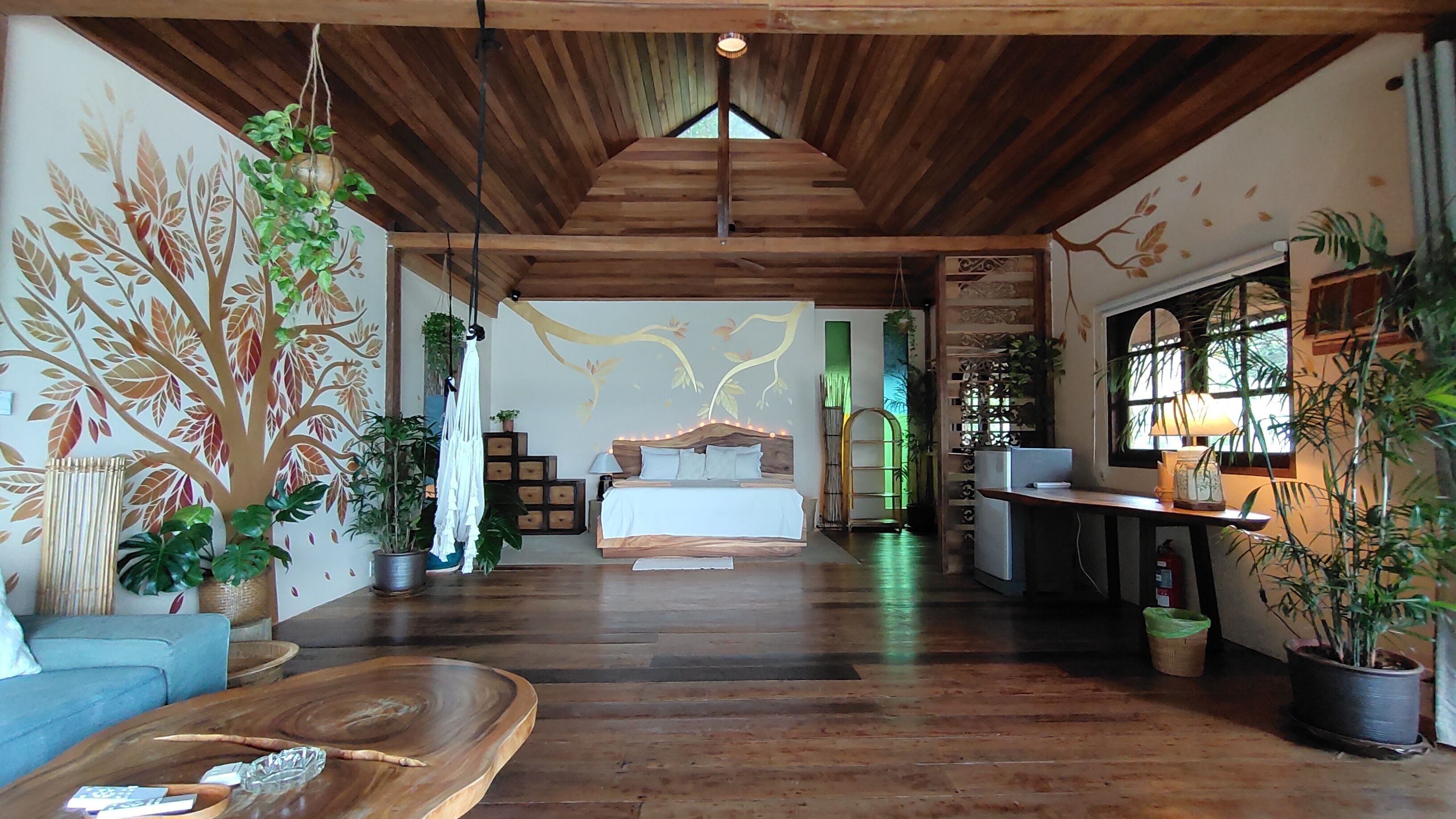 A spacious room with a wooden ceiling, a bed with a wooden headboard, a blue sofa, and various potted plants.