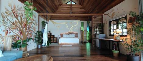 A spacious room with a wooden ceiling, a bed with a wooden headboard, a blue sofa, and various potted plants.