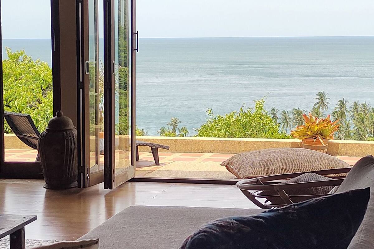 A room with a view of the ocean, a sliding glass door, a lounge chair, and a small table.