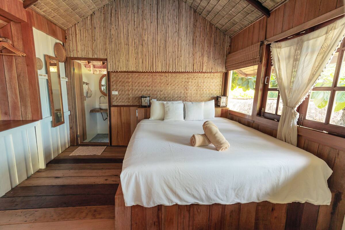 A wooden room with a large bed, a mirror, and a window with curtains.