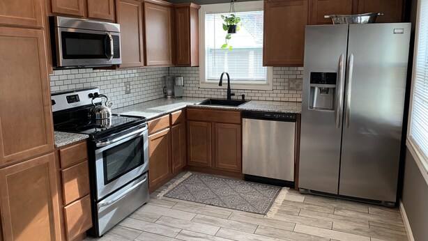 A modern kitchen with wooden cabinets, stainless steel appliances, a window with a plant, and a patterned rug.