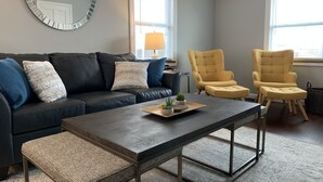 A modern living room with a dark leather sofa, two yellow armchairs, a round mirror, and a coffee table with a tray of plants.