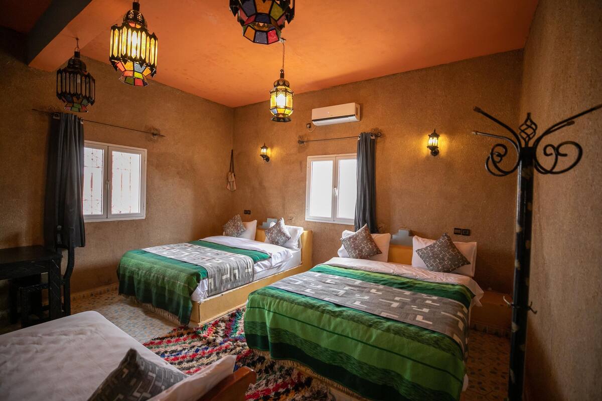 Une chambre avec deux lits, un bureau et une chaise. Elle comprend deux fenêtres avec des rideaux, un climatiseur fixé au mur et un luminaire décoratif en métal.