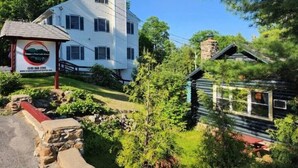 A two-story white building with a chimney, surrounded by lush greenery and a small wooden structure. - Lake George 6 Bedroom Home lake access (Lake George)