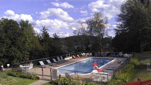 Una piscina rectangular con área cercada, rodeada de sillas de descanso y una zona verde con árboles.