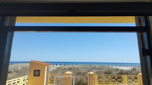 Une vue à travers une fenêtre à cadre jaune, donnant sur une plage avec une clôture et un petit bâtiment.