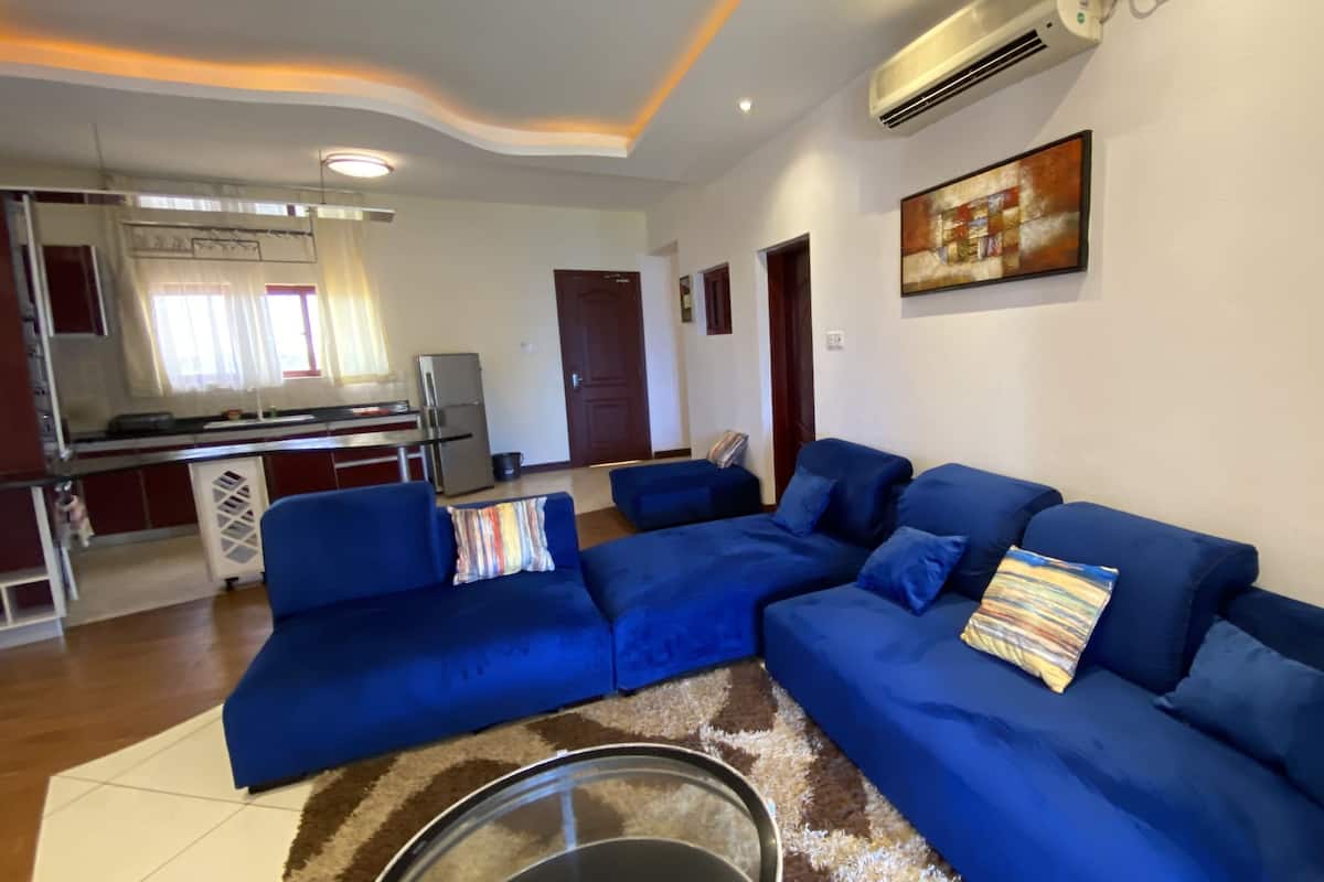 A living room with a blue sectional sofa, a kitchen area in the background, and a ceiling with recessed lighting.