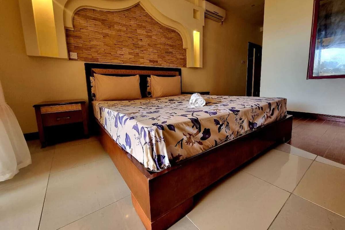 A bedroom with a large bed, a nightstand, a wooden headboard, and a wall-mounted air conditioning unit.