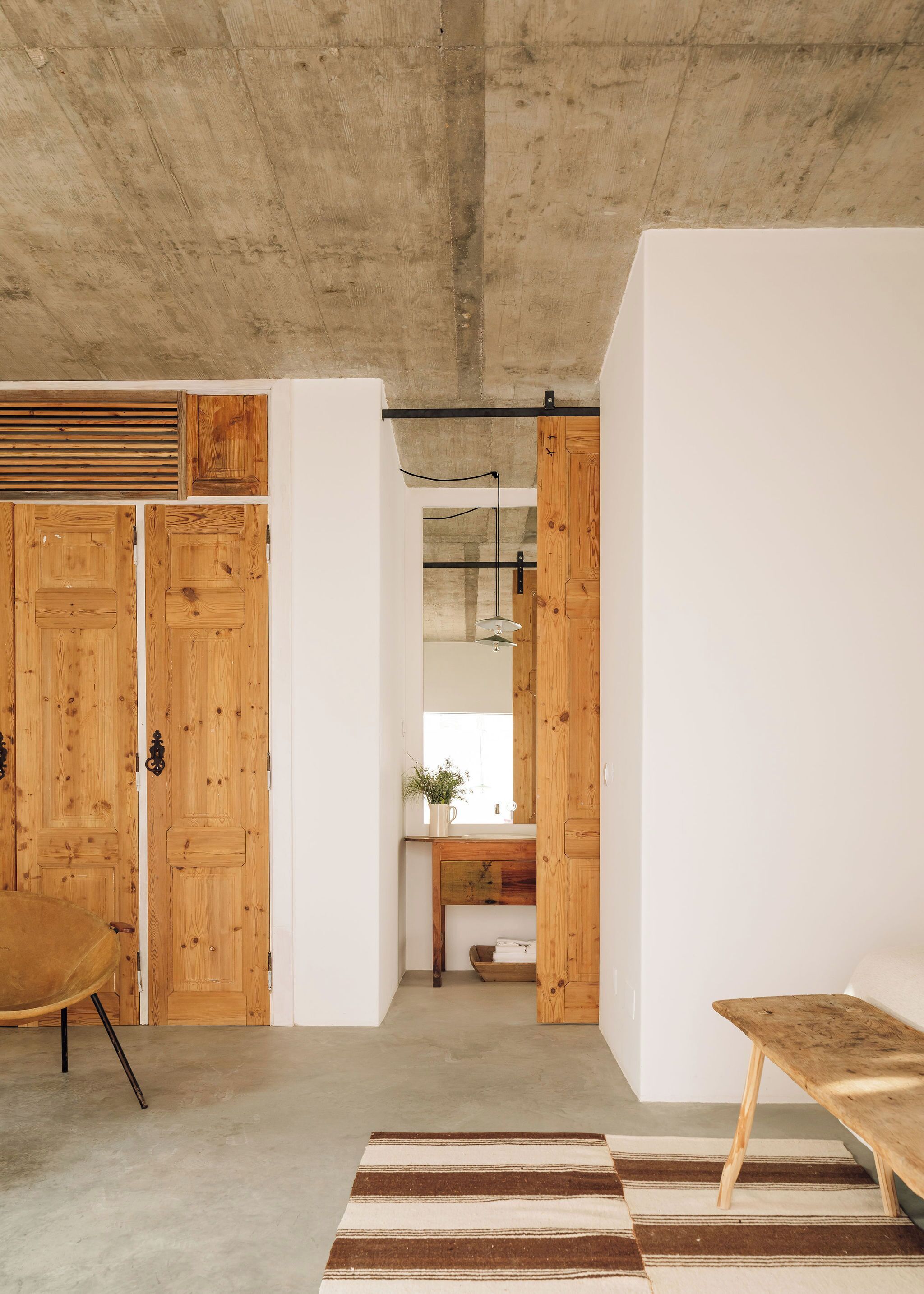 A room with wooden doors, a concrete ceiling, a striped rug, and a wooden bench.