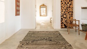 A spacious hallway with a high ceiling, a patterned rug, a wooden chair, and a stack of firewood. - Hospedaria (Tavira)