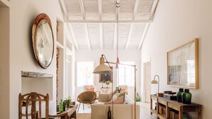 A spacious room with a high ceiling, a wooden chair, a sofa, a table with a lamp, and a painting on the wall. - Hospedaria (Tavira)
