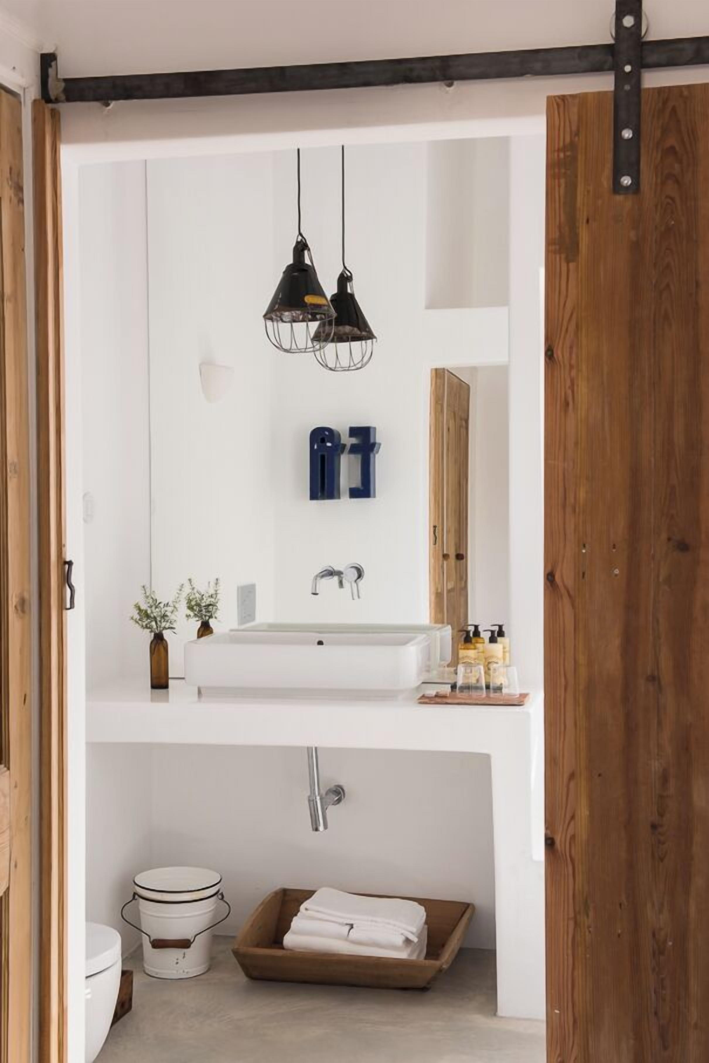 A modern bathroom with a white sink, two pendant lights, a wooden door, and a small plant.