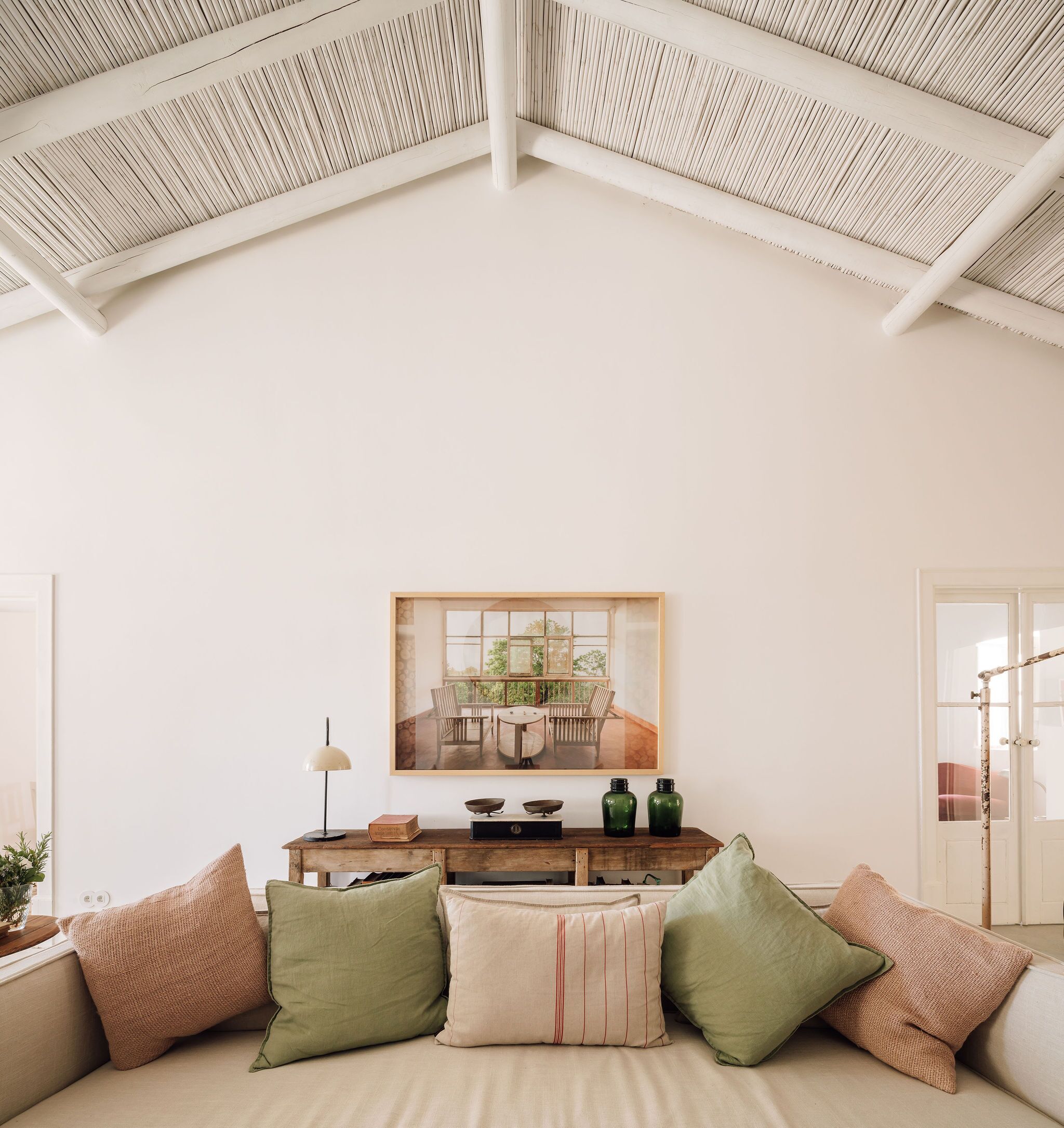 A room with a high ceiling, a wooden console table, a framed painting, and a sofa with cushions.