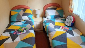 Ein Zimmer mit zwei Betten, jedes mit einer Bettdecke in geometrischem Muster in Blau, Gelb und Grau. An einem Bett befindet sich ein Nachttisch mit einer Lampe und einem Buch.