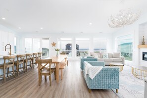A spacious living area with a wooden dining table, blue upholstered sofa, and a view of the ocean through large windows. - Beautiful 6 bedroom beach house with pool and elevator! Steps from the beach! (Oak Island)
